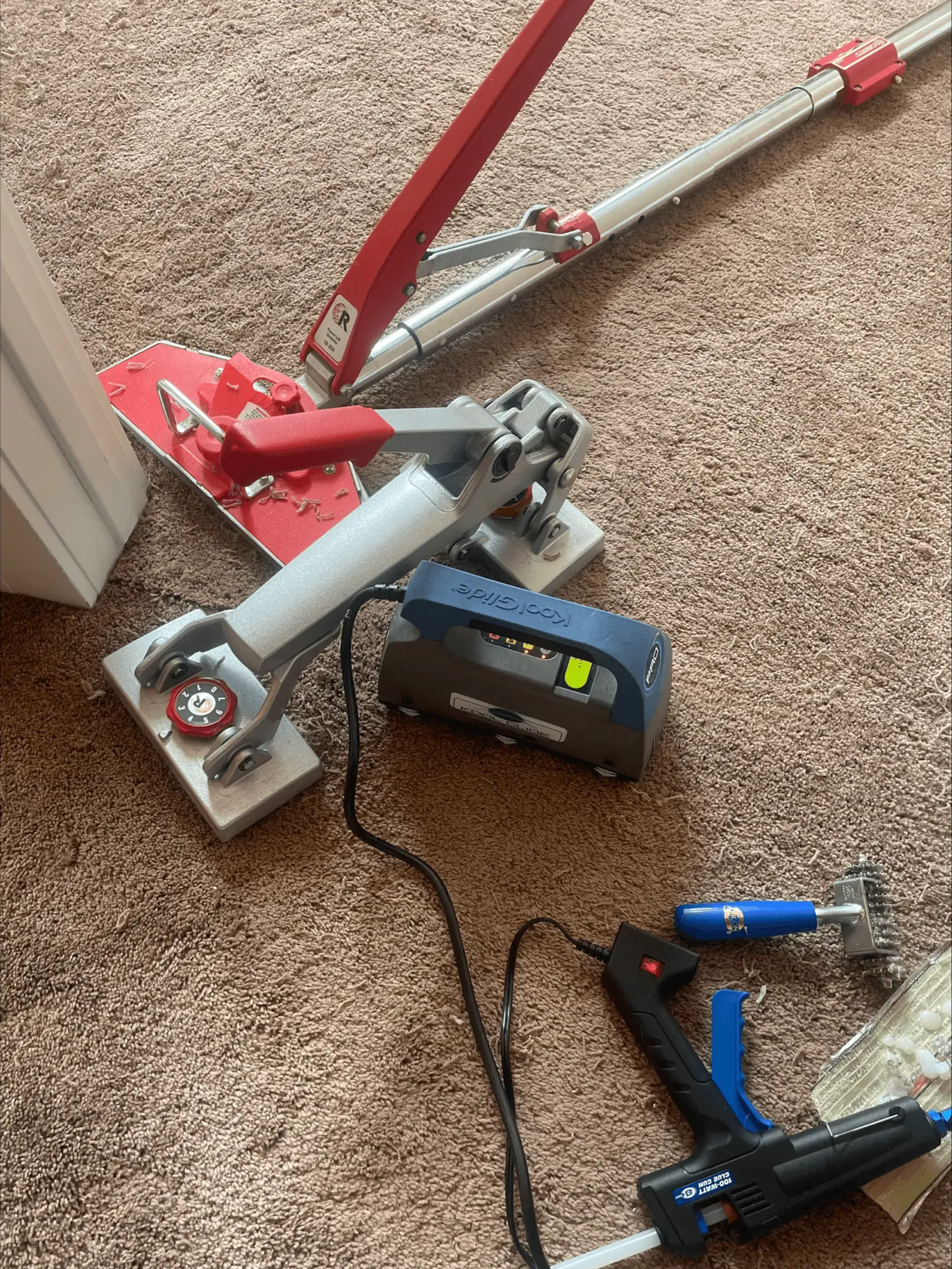 Professional carpet repair tools laid out on beige carpet — power stretcher, seam iron, glue gun, and tackless tools