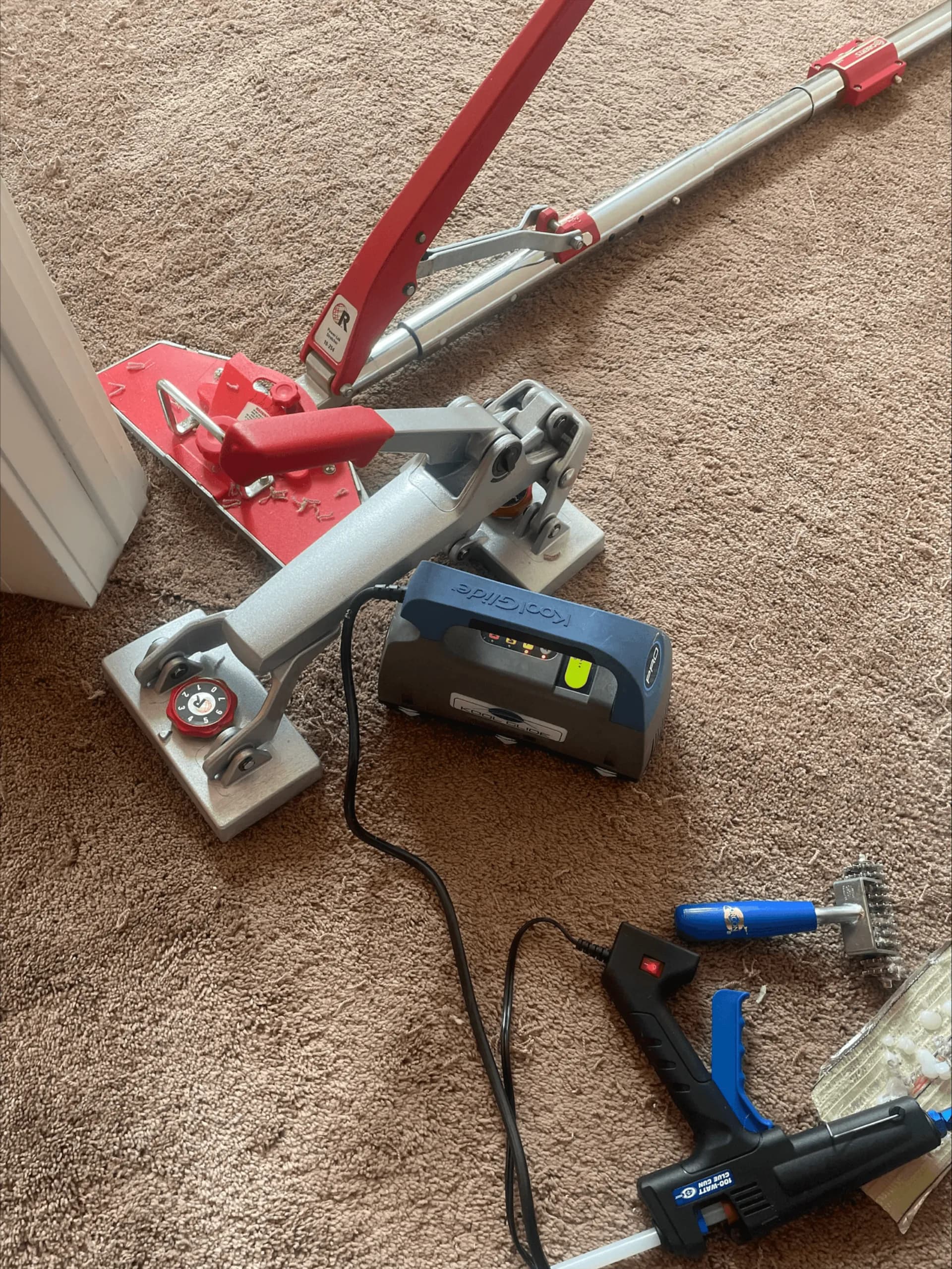 Professional carpet repair tools laid out on beige carpet — power stretcher, seam iron, glue gun, and tackless tools